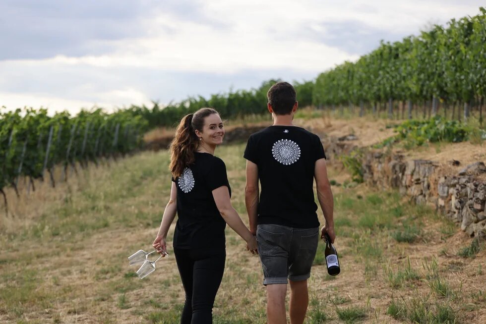 Zwei Personen laufen durch Weinberge als Rückenfiguren. Die linke Person dreht sich um und schaut lächelnd in die Kamera. 