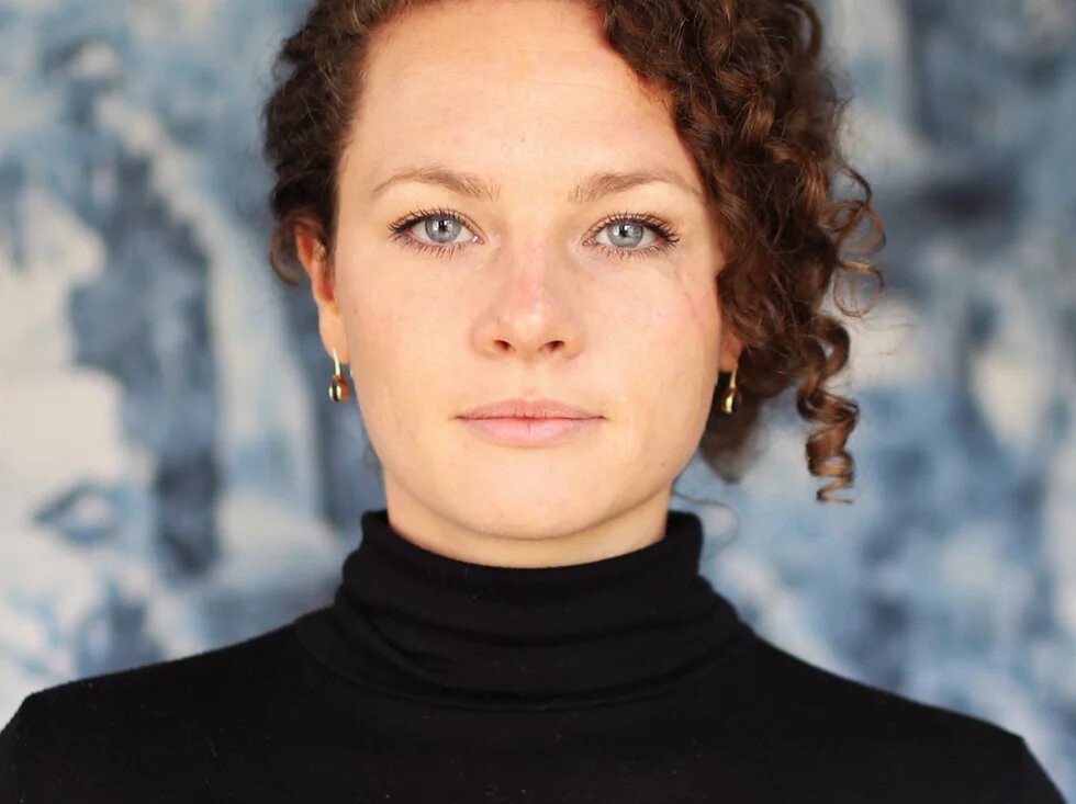 Eine weiblich gelesene Person sieht mit blaugrauen Augen direkt die betrachtende Person an. Sie hat braune gelockte Haare, die sie hochgesteckt trägt. Der Gesichtsausdruck ist ruhig und selbstsicher.