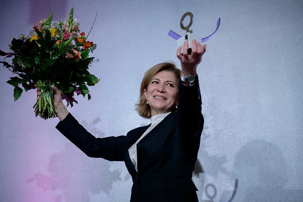 Joumana Seif hält den Anne-Klein-Frauenpreis in einer Hand und einen Blumenstrauß in der anderen. Sie lächelt vor einem neutralen Hintergrund.