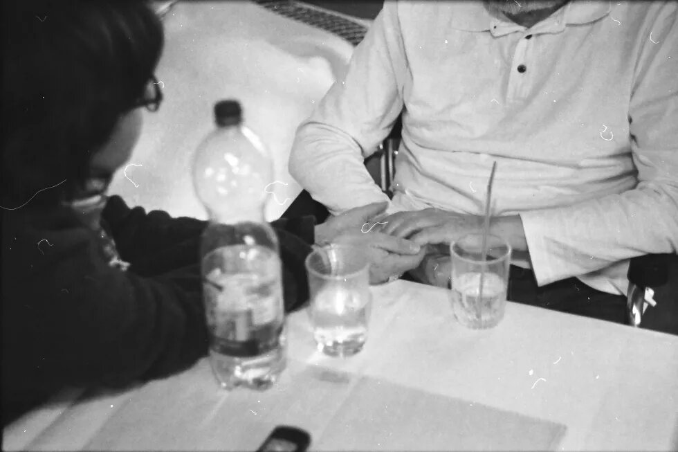 „Rund um die Uhr für Sie da.“ Frau mittleren Alters hält die Hand eines alten Menschen, im Vordergrund Wasserflasche und zwei Gläser
