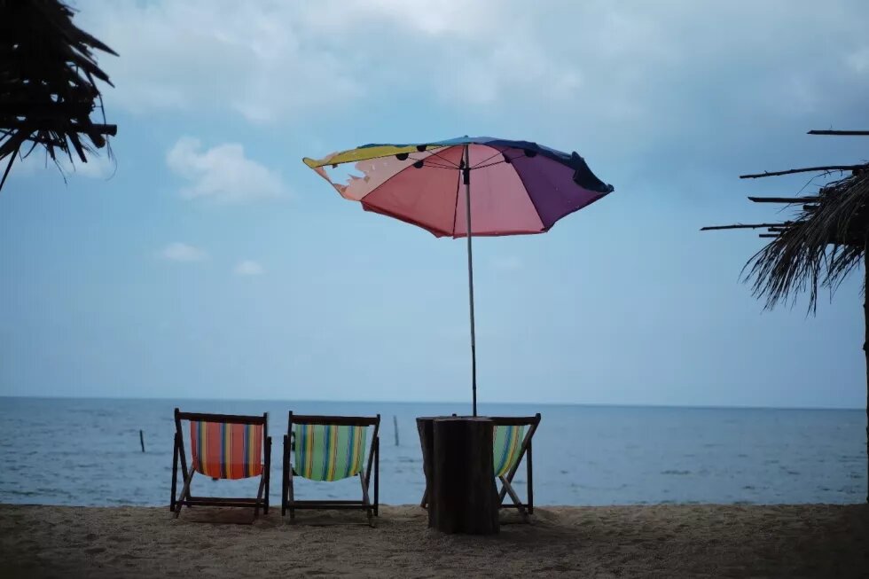 Foto an einem Strand. Im Bild sind drei Liegestühle und ein Sonnenschimr. Am Bildraund stehen Palmen.