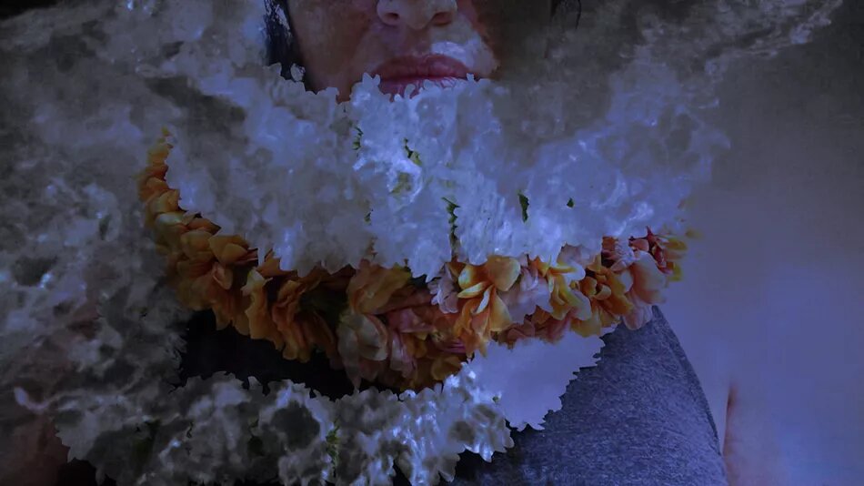 Video Still: Together Apart Eine Person mit Blumenkränzen um den Hals