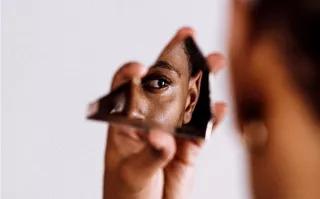 reflection of PoC/black woman looking into a piece of a mirror she holds