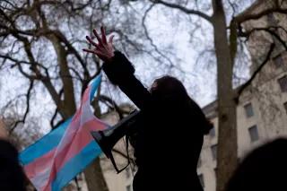 Trans Rights Protest London, April 2022 Eine Person hält ein Megafon, Im Hintergrund Bäume und Häuser, links neben der Person weht eine trans Flagge