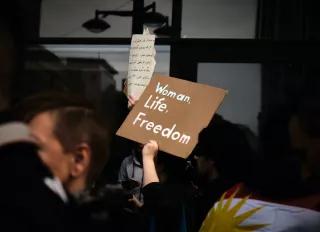 Auf dem Foto sieht man Protestierende gegen das Regime im Iran. Im Hintergrund ist ein Plakat mit Schriftzeichen zu sehen, die aber unscharf sind. Im Vordergrund hält eine Hand ein Plakat mit der Aufschrift Women Life Freedom hoch. Im unteren rechten Bildrand ist ein Ausschnitt einer Iranflagge abgebildet. 