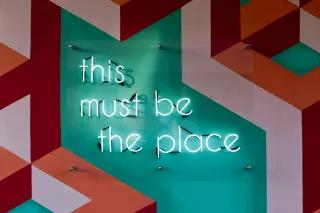 This must be the place Foto einer bunten Wand auf der eine Neonleuchtschrift angebracht ist, die besagt: this must be the place