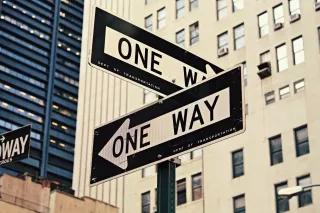 Foto einer Kreuzung in New York. Zwei Straßenschilder mit "One Way"-Aufschrift weisen in verschiedene Richtungen. 