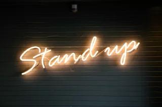stand up Leuchtzeichen mit der Schrift "Stand up" frontal fotografiert vor dunklem Hintergrund