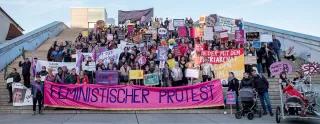 Feministischer Protest Eine Gruppe von circa 200 Demonstrierenden steht auf einer Treppe im Freien für ein Gruppenfoto. Sie tragen Protestschilder und Plakate. Im Fordergrund tragen Leute ein Banner mit der Aufschrift: Feministischer Protest