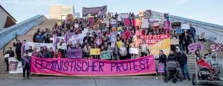 Eine Gruppe von circa 200 Demonstrierenden steht auf einer Treppe im Freien für ein Gruppenfoto. Sie tragen Protestschilder und Plakate. Im Fordergrund tragen Leute ein Banner mit der Aufschrift: Feministischer Protest