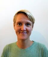Eine junge weisse Person mit kurzen blonden Haaren und ovalem Gesicht, lächelt offen in die Kamera. Sie ist etwa mitte dreißig, trägt ein hellgrünes Shirt und silberne Ohrpiercings.