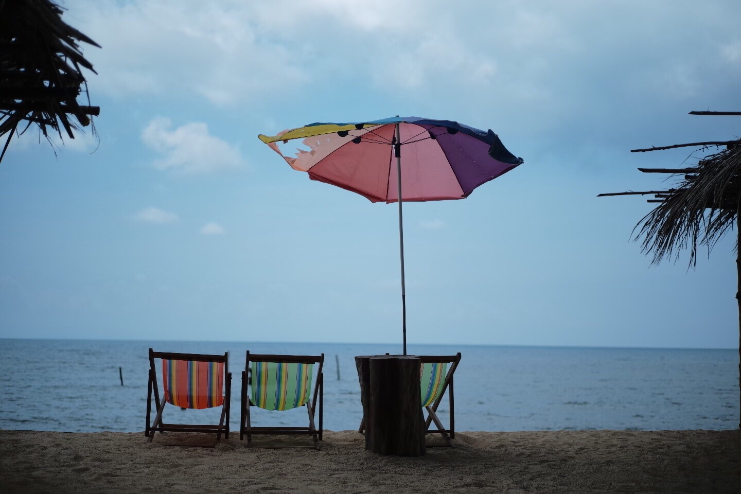 Foto an einem Strand. Im Bild sind drei Liegestühle und ein Sonnenschimr. Am Bildraund stehen Palmen.