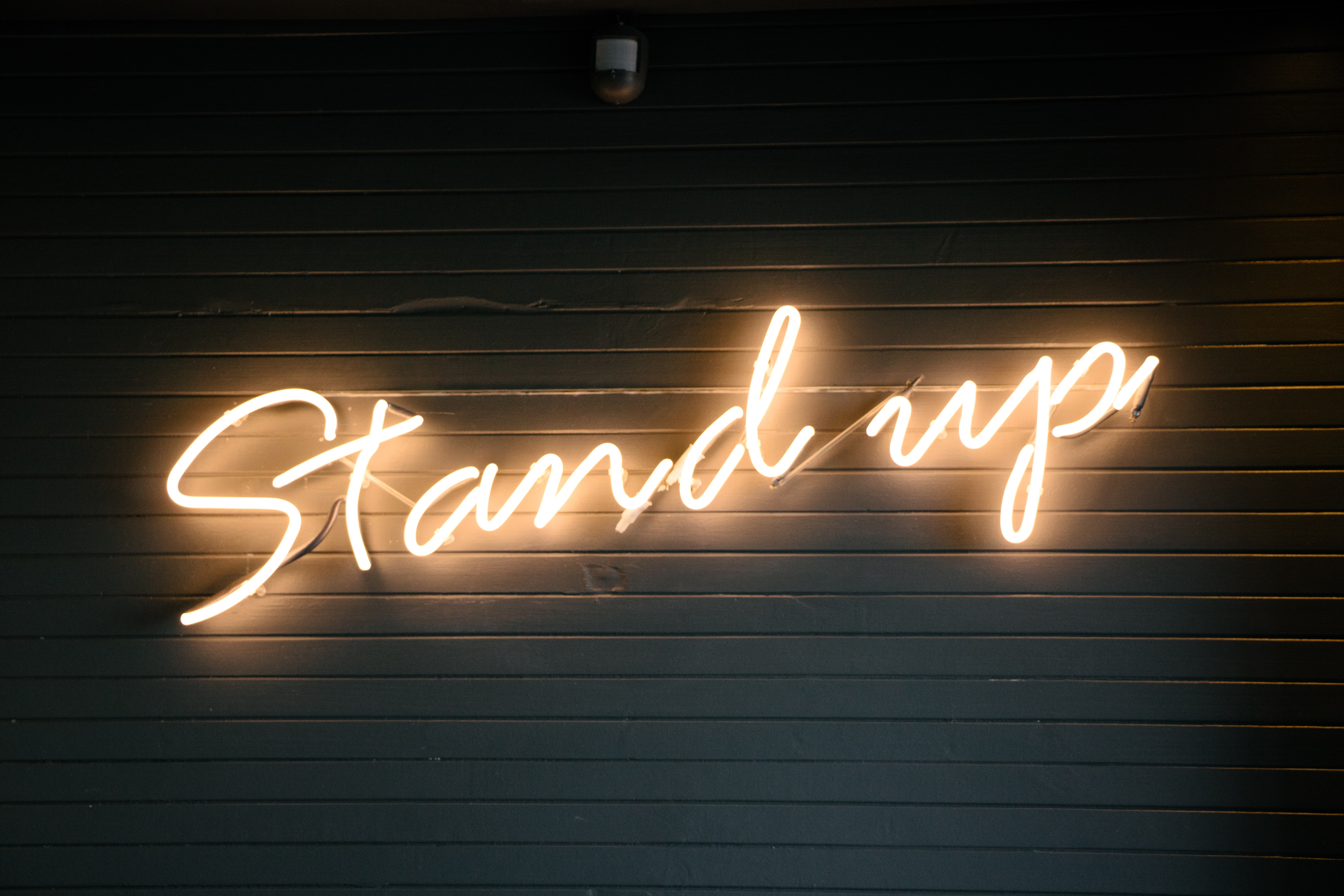 stand up Leuchtzeichen mit der Schrift "Stand up" frontal fotografiert vor dunklem Hintergrund