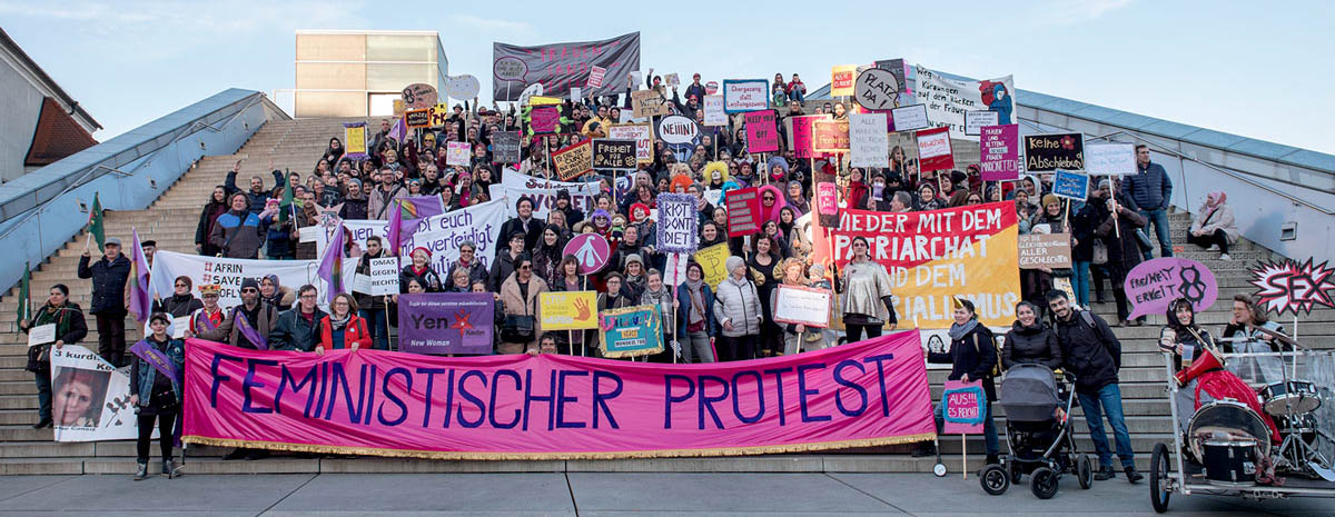 Feministischer Protest Eine Gruppe von circa 200 Demonstrierenden steht auf einer Treppe im Freien für ein Gruppenfoto. Sie tragen Protestschilder und Plakate. Im Fordergrund tragen Leute ein Banner mit der Aufschrift: Feministischer Protest