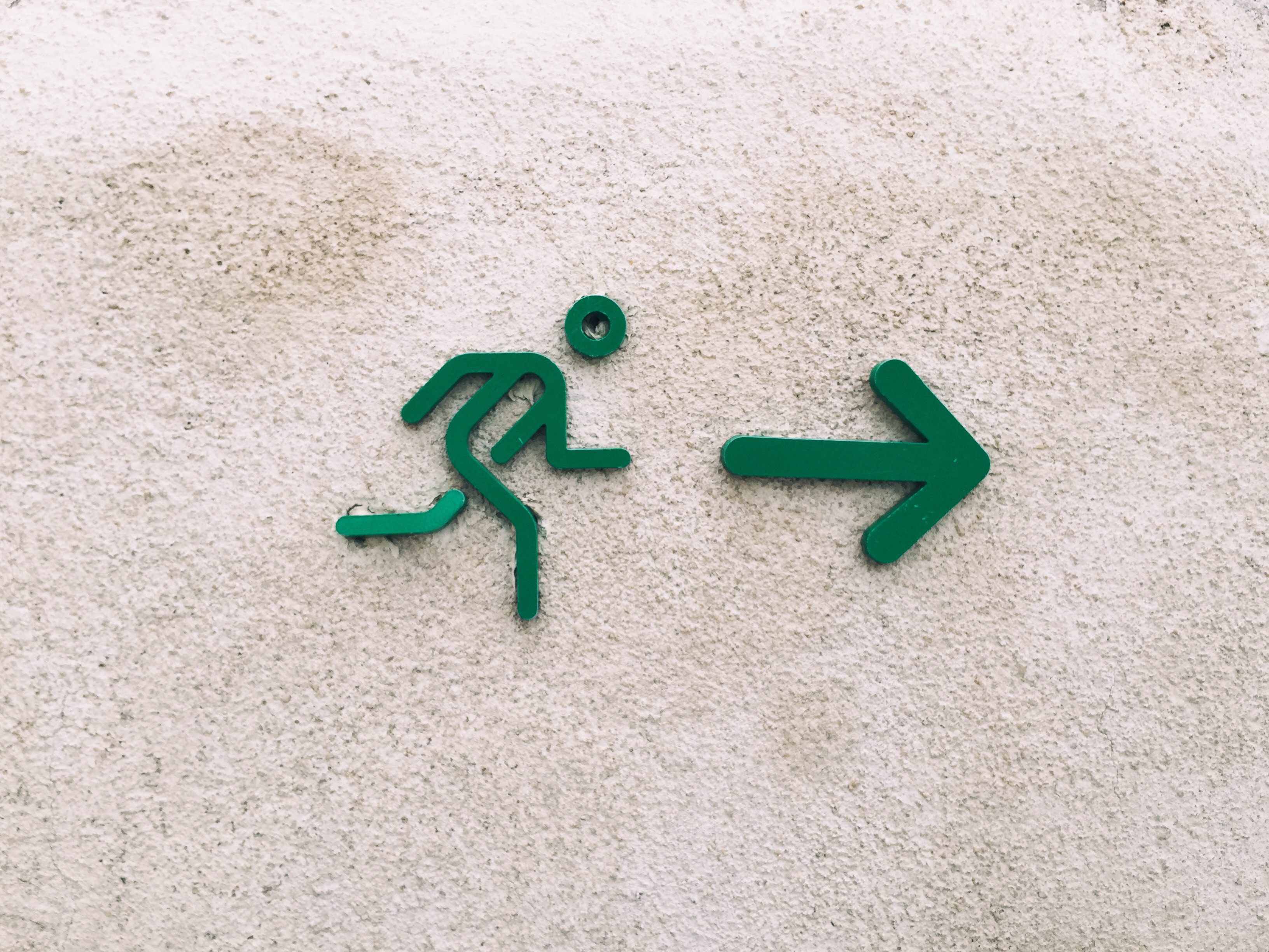 Auf einer hellgrauen Hauswand ist ein "exit"-Schild angebract (Eine Person läuft in Richtung eines Pfeils)